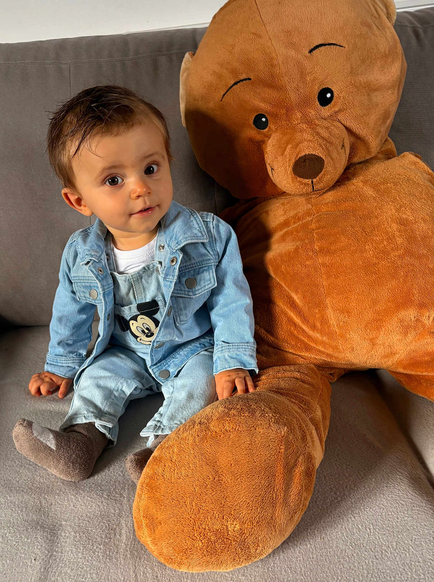 Julien a rejoint le concours — aidez-le/la à gagner de superbes lots ! toddler, child, denim_jacket, denim_overalls, socks, plush_toy, teddy_bear, couch, indoor, cute, smiling, face, person, seated, soft_toy, home, casual_clothing, brown, gray, fabric