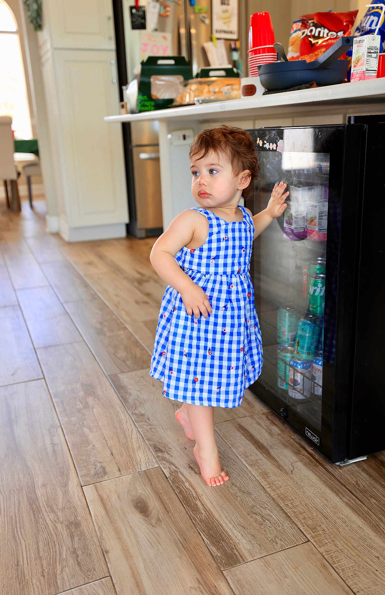 Raea joined the competition — help win amazing prizes! toddler, child, blue_dress, barefoot, kitchen, wooden_floor, fridge, glass_door, beverage_cans, snacks, indoor, curly_hair, exploring, standing, person, casual_clothing, daylight, household, interior, young_child