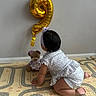 baby, balloon, barefoot, child, crawling, crawling_baby, crocheted_teddy_bear, cute, floor_mat, gold_balloon, headband, home, indoor, infant, number_balloon, patterned_clothing, play_mat, teddy_bear, toy, wall