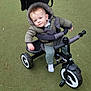 baby, child, curious, daylight, footwear, green_surface, handlebar, hood, jacket, nature, outdoor, park, person, playground, stroller, toddler, toy, tricycle, wheel, winter_clothing
