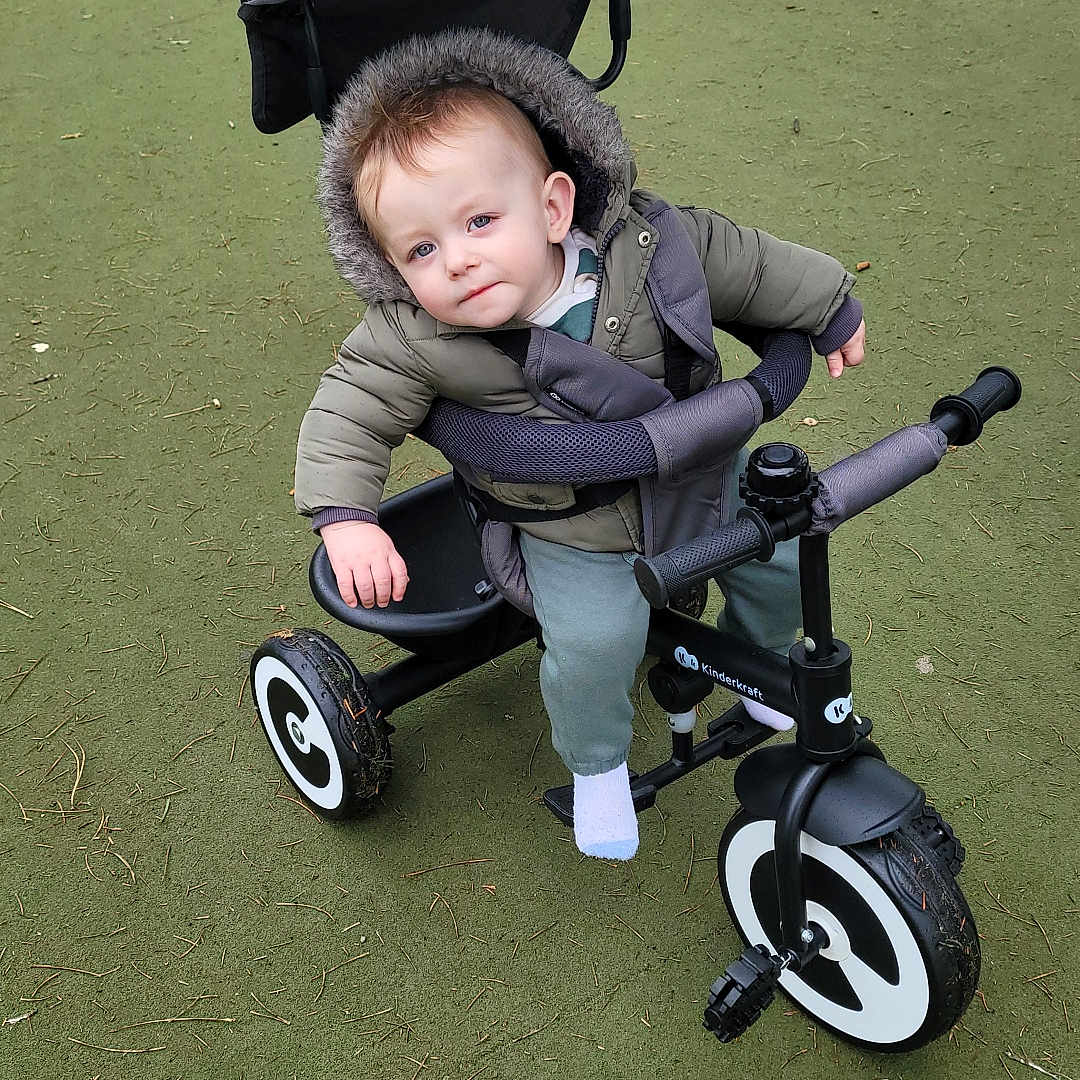 Ollie joined the competition — help win amazing prizes! baby, child, curious, daylight, footwear, green_surface, handlebar, hood, jacket, nature, outdoor, park, person, playground, stroller, toddler, toy, tricycle, wheel, winter_clothing