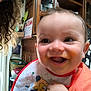 baby, smiling, indoor, bib, orange_clothing, curly_hair, wooden_furniture, smile, happy, face, person, child, messy_face, cabinet, basket, radio, chair, home, closeup, portrait