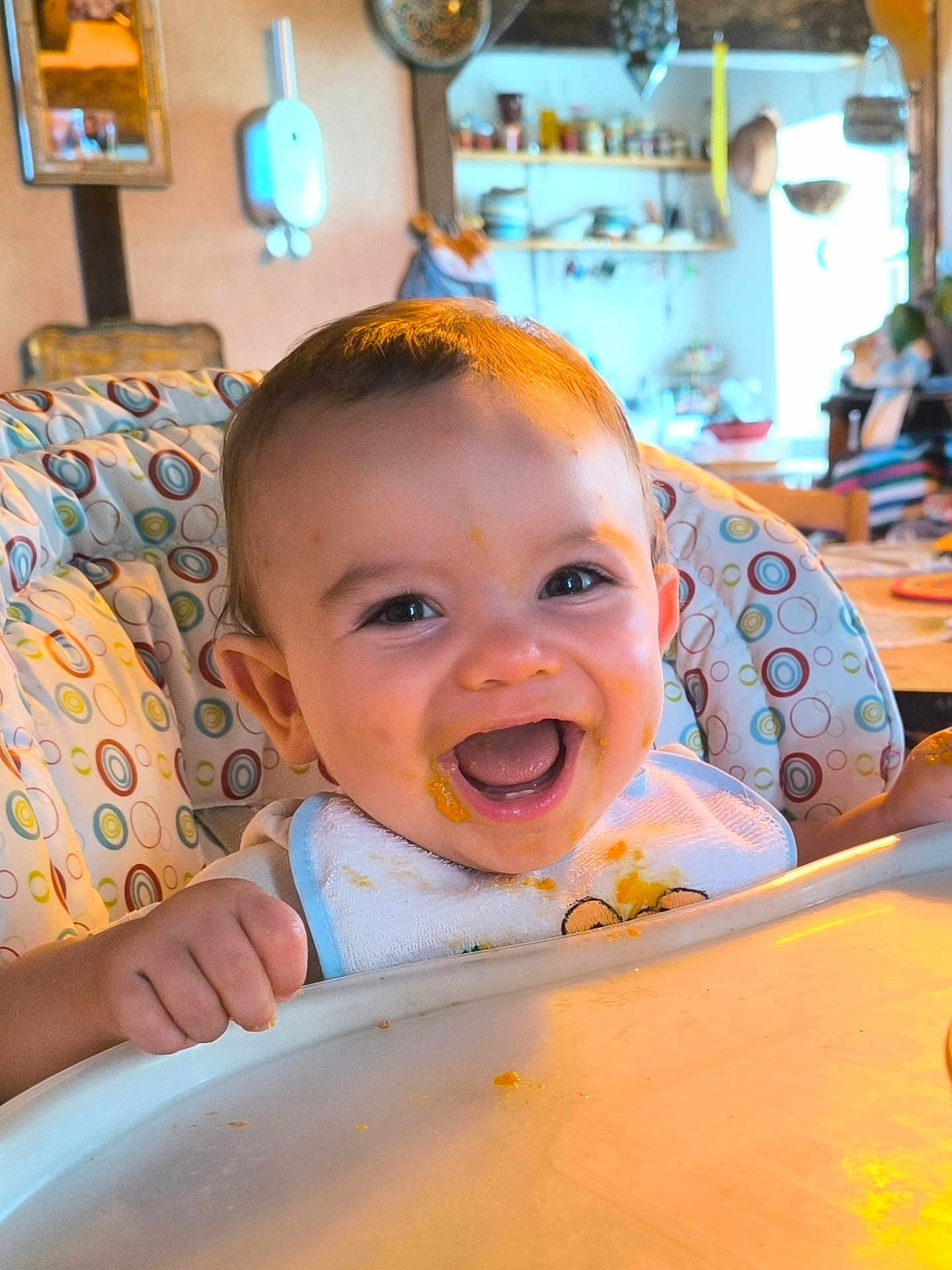 Louison participe au concours pour gagner de l'argent avec cette photo : baby, high_chair, food, bib, smiling, happy, face, child, indoor, kitchen, furniture, person, messy, feeding, cute, mouth_open, table, colorful, infant, dining