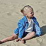 child, blonde_hair, pony_tail, denim_jacket, white_top, sand, barefoot, sitting, outdoor, sunlight, shadow, casual_clothing, young_child, curious_expression, legs, hand, summer, playful, nature, daytime