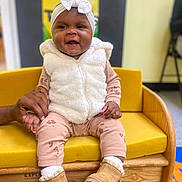 Journei is registered to the contest to win money with this photo: baby, child, smiling, sitting, yellow_sofa, white_vest, headband, bow, boots, hand_holding, colorful_background, indoor, cute, happy, clothing, wooden_furniture, person, playroom, carpet, young_child