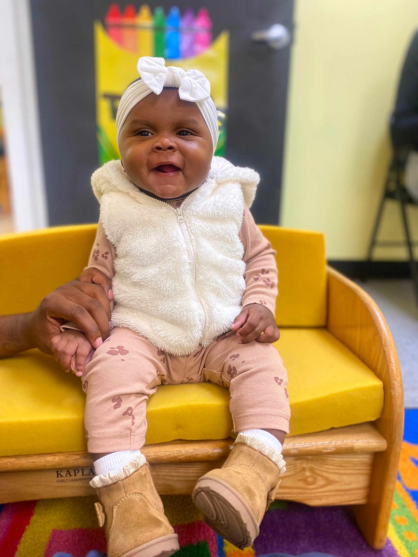Journei is registered to the contest to win money with this photo: baby, child, smiling, sitting, yellow_sofa, white_vest, headband, bow, boots, hand_holding, colorful_background, indoor, cute, happy, clothing, wooden_furniture, person, playroom, carpet, young_child