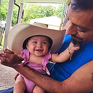 Victoria is registered to the contest to win money with this photo: baby, child, man, cowboy_hat, smile, holding, porch, outdoor, trees, tattoo, blue_shirt, happy, infant, playful, bracelet, person, nature, casual, family, bonding