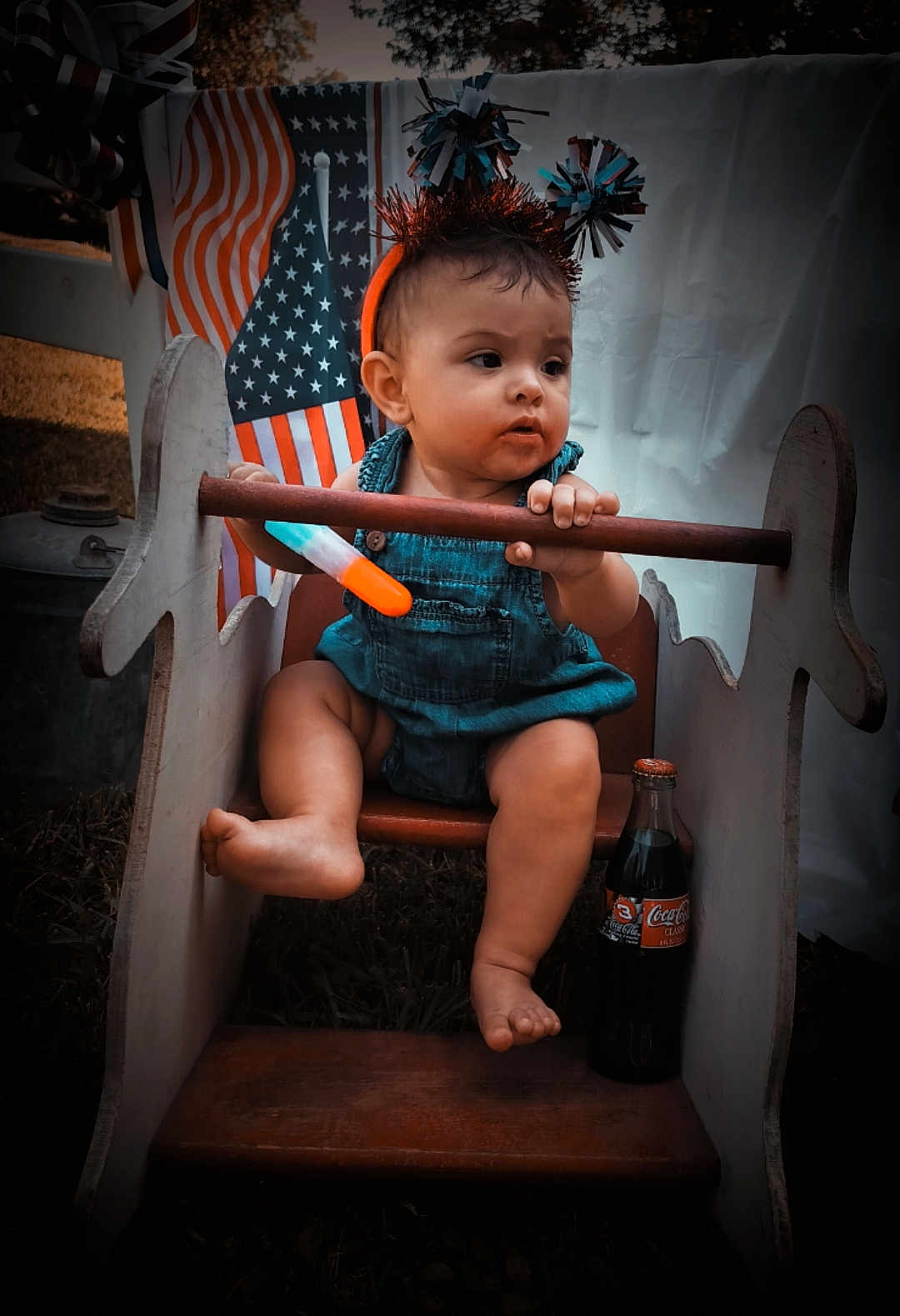 Victoria is registered to the contest to win money with this photo: baby, child, rocking_horse, wooden_toy, american_flag, headgear, outdoor, grass, bottle, coca_cola, drink, summer, portrait, cute, festive, holiday, play, seat, curious, sitting