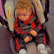 Zarek is registered to the contest to win money with this photo: child, toddler, baby, curly_hair, car_seat, safety_harness, seated, red_clothing, black_clothing, tracksuit, socks, indoor, floor, play_area, portrait, looking_away, serious_expression, vehicle_seat, sparkles, photography
