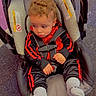child, toddler, baby, curly_hair, car_seat, safety_harness, seated, red_clothing, black_clothing, tracksuit, socks, indoor, floor, play_area, portrait, looking_away, serious_expression, vehicle_seat, sparkles, photography