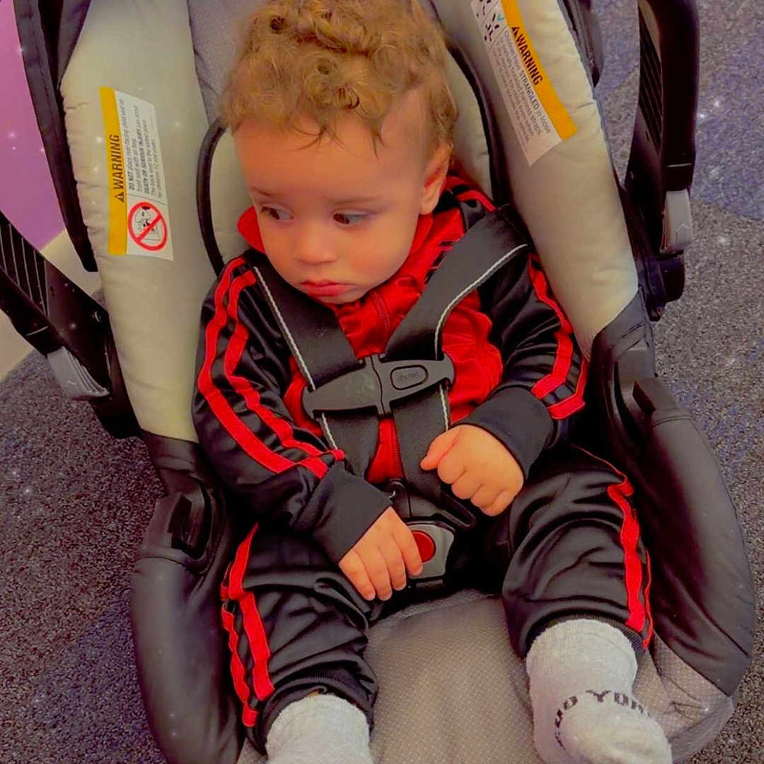 Zarek is registered to the contest to win money with this photo: child, toddler, baby, curly_hair, car_seat, safety_harness, seated, red_clothing, black_clothing, tracksuit, socks, indoor, floor, play_area, portrait, looking_away, serious_expression, vehicle_seat, sparkles, photography