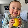 baby, child, smile, apple, hands, face, indoor, clothing, table, water_bottle, television, clock, chair, furniture, happy, cute, skin, eyelashes, home, person