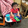 baby, child, infant, smiling, sitting, cap, hat, red_shirt, table, colorful, superhero, toy, cup, indoor, store, shelving, shoes, fabric, portrait, happy
