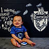 baby, child, smiling, blue_clothing, onesie, sitting, footprints, harry_potter, ravenclaw, crest, wall_art, indoor, happy, cute, infant, portrait, playful, person, floor, background