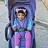 baby, child, clothing, curly_hair, daylight, denim_jacket, happy, jogging_pants, outdoor, person, pink_headband, purple_stroller, seatbelt, sidewalk, smiling, stroller, toddler, urban, walking, white_sneakers