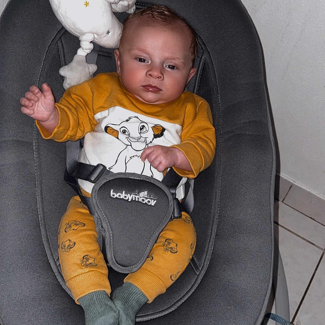 Tiago participe au concours pour gagner de l'argent avec cette photo : baby, child, infant, baby_rocker, seat, mustard_outfit, cartoon_print, plush_toy, moon_shaped, teddy_bear, gray_fabric, floor_tiles, straps, socks, indoor, serious_expression, baby_clothes, baby_accessory, baby_seat, baby_gadget