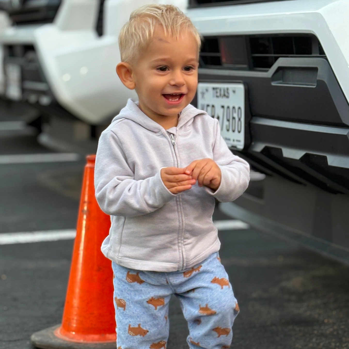 Poyraz is registered to the contest to win money with this photo: animal_print, blonde_hair, casual_clothing, child, cute, daytime, happy, hoodie, outdoor, pants, parking_lot, person, playful, smiling, sneakers, standing, toddler, traffic_cone, vehicle, white_car