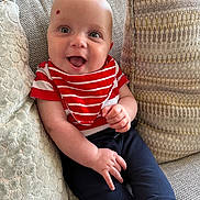 George joined the competition — help win amazing prizes! baby, smiling, couch, red_striped_bib, navy_pants, infant, happy, sitting, indoor, face, skin, child, clothing, cute, portrait, cozy, cushion, home, person, young_child