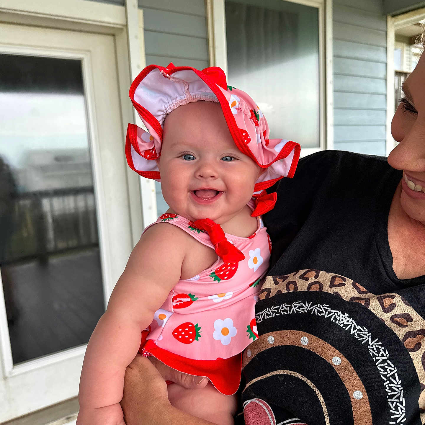Avery is registered to the contest to win money with this photo: adult, arm, baby, child, clothing, door, face, flower, happy, holding, outdoor, pattern, person, porch, skin, smiling, strawberry, sunhat, swimsuit, window
