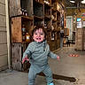 child, toddler, smiling, happy, standing, portrait, indoor, store, cafe, rustic, wooden_crates, jars, shelves, food_products, clothing, gray_outfit, blue_shoes, cartoon_shoes, floor, lighting