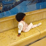 Louna participe au concours pour gagner de l'argent avec cette photo : bench, casual_clothing, child, cityscape, curly_hair, floor, glass, indoor, jacket, marble, person, portrait, reflection, relaxed, seated, side_view, sneakers, urban, wall, young
