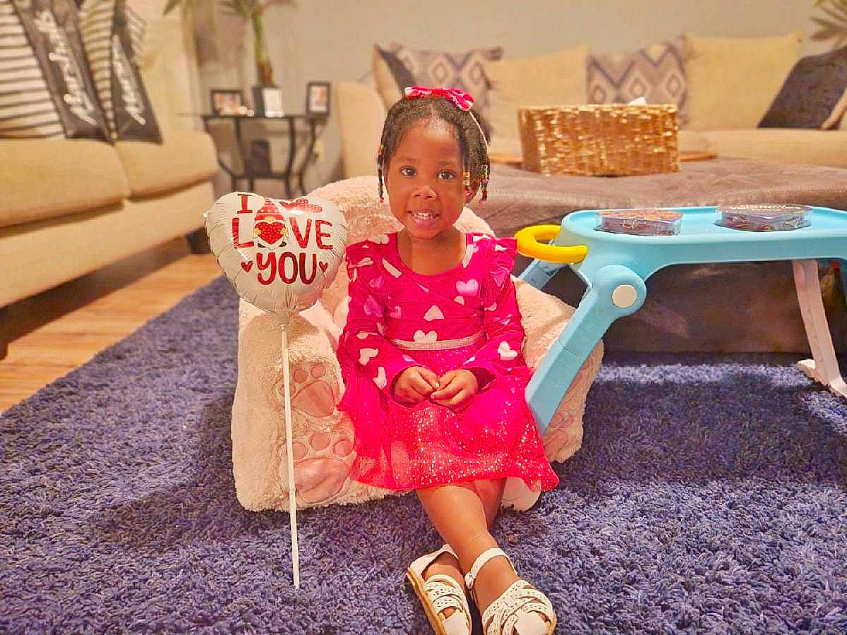 Zyana is registered to the contest to win money with this photo: baby_toddler_clothing, couch, dress, event, fashion_design, floor, flooring, fun, furniture, happy, human_leg, leg, leisure, magenta, person, pink, sandal, sitting, smile, table