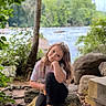 casual_clothing, child, curly_hair, daylight, footwear, forest, girl, greenery, happy, leaves, nature, outdoor, portrait, relaxing, river, rock, scenic, sitting, smile, tree