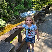 Rilynn-marie is registered to the contest to win money with this photo: child, girl, outdoor, river, wooden_railing, stone_path, greenery, trees, sunlight, smiling, casual_clothing, shorts, tshirt, sneakers, socks, backpack, nature, daylight, happy, portrait