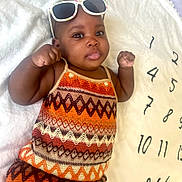 Rihanna Jemila participe au concours pour gagner de l'argent avec cette photo : baby, blanket, child, closeup, colorful_clothing, curious, cute, expression, fashion, headwear, indoors, infant, lying_down, numbers, patterned_outfit, playful, portrait, raised_fists, soft_texture, sunglasses