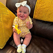 Holly joined the competition — help win amazing prizes! baby, child, smiling, yellow_clothing, headband, bow, toy, airplane_toy, socks, couch, leather, cushions, yellow_cushions, happy, indoor, person, infant, cute, portrait, seated