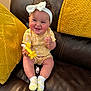 baby, child, smiling, yellow_clothing, headband, bow, toy, airplane_toy, socks, couch, leather, cushions, yellow_cushions, happy, indoor, person, infant, cute, portrait, seated