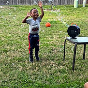 Amari is registered to the contest to win money with this photo: child, bubbles, bubble_machine, jumping, grass, backyard, basketball, table, fence, happy, outdoor, toy, play, person, smiling, casual_clothing, sneakers, daylight, greenery, fun