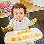 Olivia is registered to the contest to win money with this photo: bib, child, curly_hair, cute, expression, floor, food, high_chair, home, indoor, letters, person, plastic_container, playtime, seated, snack, toddler, toys, tray, yellow_clothing