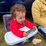 Olivia is registered to the contest to win money with this photo: bright_starts, casual_clothing, child, curly_hair, daylight, feet, grass, happy, high_chair, outdoor, person, playful, red_sweater, reflection, sitting, smartphone, sock, toddler, toy, yellow_hoodie