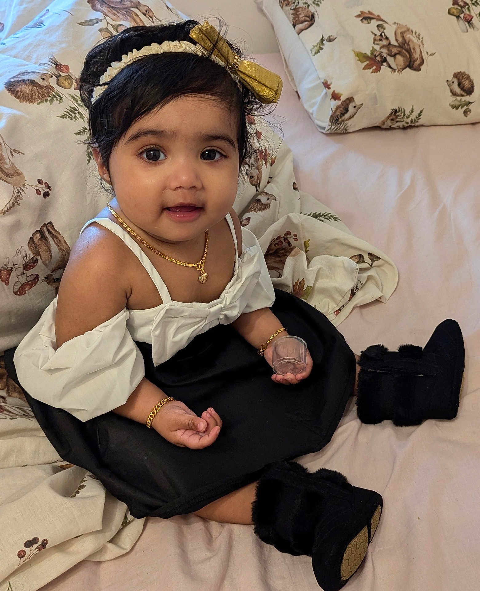 Hanika is registered to the contest to win money with this photo: baby, child, girl, black_hair, headband, gold_jewelry, dress, boots, bed, pillow, blanket, animal_print, indoor, smiling, cute, sitting, person, portrait, hand, curious