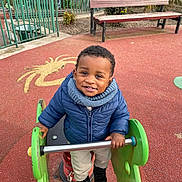 Ilyan a rejoint le concours — aidez-le/la à gagner de superbes lots ! bench, child, clothing, fence, footwear, greenery, happy, jacket, metal, outdoor, park, person, play_area, playground, rubber_flooring, scarf, smile, spring_rider, toddler, toy
