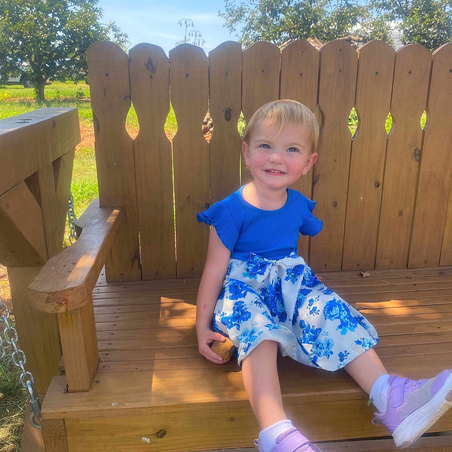 Rosie joined the competition — help win amazing prizes! child, toddler, girl, smiling, outdoor, swing, wooden_fence, backyard, garden, sunlight, blue_shirt, floral_dress, purple_shoes, blond_hair, portrait, seated, playground, happy, summer, family_photo