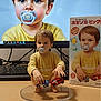 figurine, toddler, pacifier, toy_cars, computer_screen, portrait, product_box, realistic_art, child_figure, yellow_shirt, plastic_stand, desktop_keyboard, table_surface, blue_eyes, brown_hair, digital_art, collectible, miniature, indoor, photography