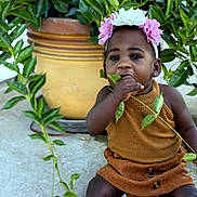 Armani is registered to the contest to win money with this photo: toddler, child, flower_headband, leaf, plant, pot, outdoor, curious, sitting, brown_clothing, nature, greenery, cute, exploring, face, portrait, young_child, headwear, plant_pot, daylight