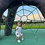 child, toddler, playground, rope_structure, green_rope, outdoor, grass, sky, clouds, sunset, pigtails, white_shirt, shorts, shoes, fence, playing, young_child, park, fun, daylight