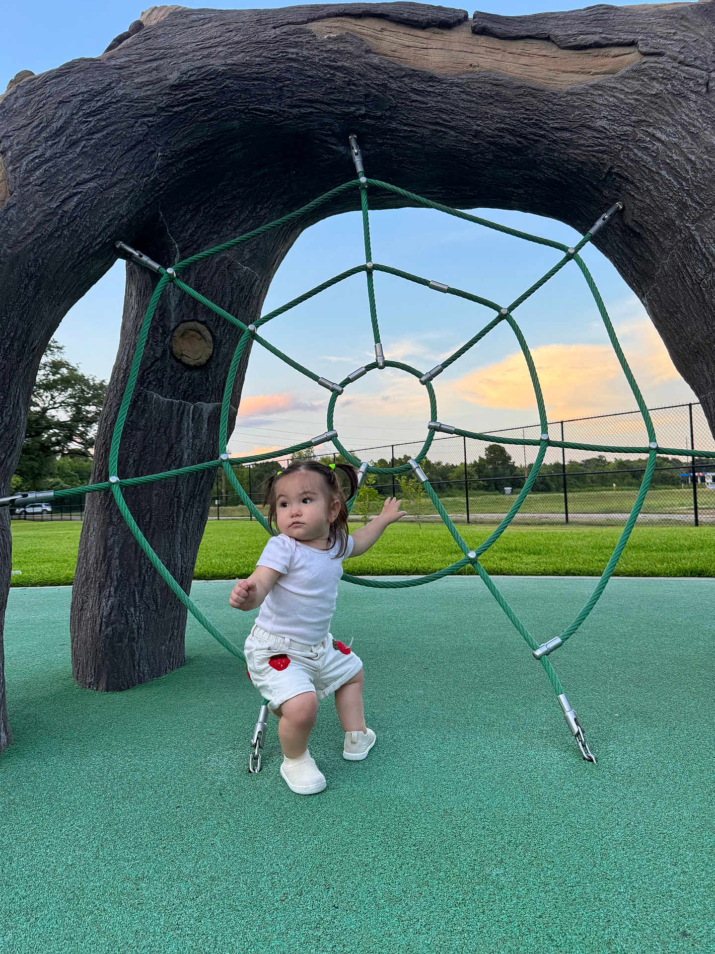 Deniz joined the competition — help win amazing prizes! child, toddler, playground, rope_structure, green_rope, outdoor, grass, sky, clouds, sunset, pigtails, white_shirt, shorts, shoes, fence, playing, young_child, park, fun, daylight