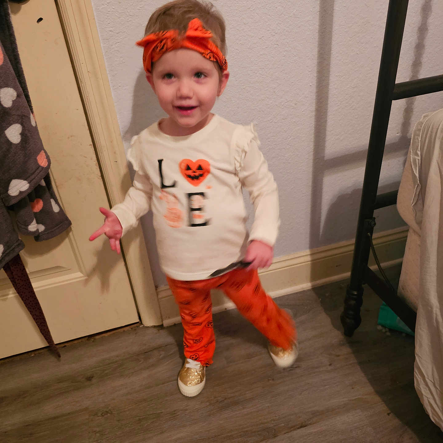 Athena is registered to the contest to win money with this photo: blur, child, clothing, door, floor, footwear, gold_shoes, happy, headband, indoor, orange, pants, person, playful, pose, pumpkin, shirt, toddler, wall, young_child