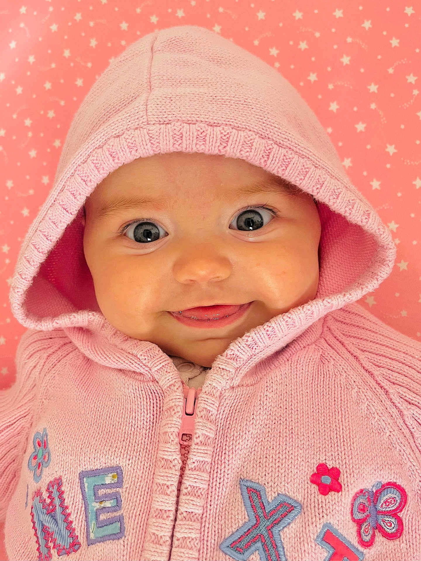 Bethany is registered to the contest to win money with this photo: baby, pink_hoodie, knitted_clothing, smiling, tongue_out, close_up, cute, infant, face, blue_eyes, child, hood, patterned_background, star_pattern, soft_texture, happy, portrait, warm_clothing, pink_color, baby_girl