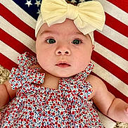 Brenna is registered to the contest to win money with this photo: baby, infant, headband, bow, dress, floral_pattern, american_flag, blanket, face, eyes, skin, arm, cute, child, portrait, laying_down, fabric, pattern, closeup, indoors