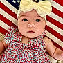baby, infant, headband, bow, dress, floral_pattern, american_flag, blanket, face, eyes, skin, arm, cute, child, portrait, laying_down, fabric, pattern, closeup, indoors