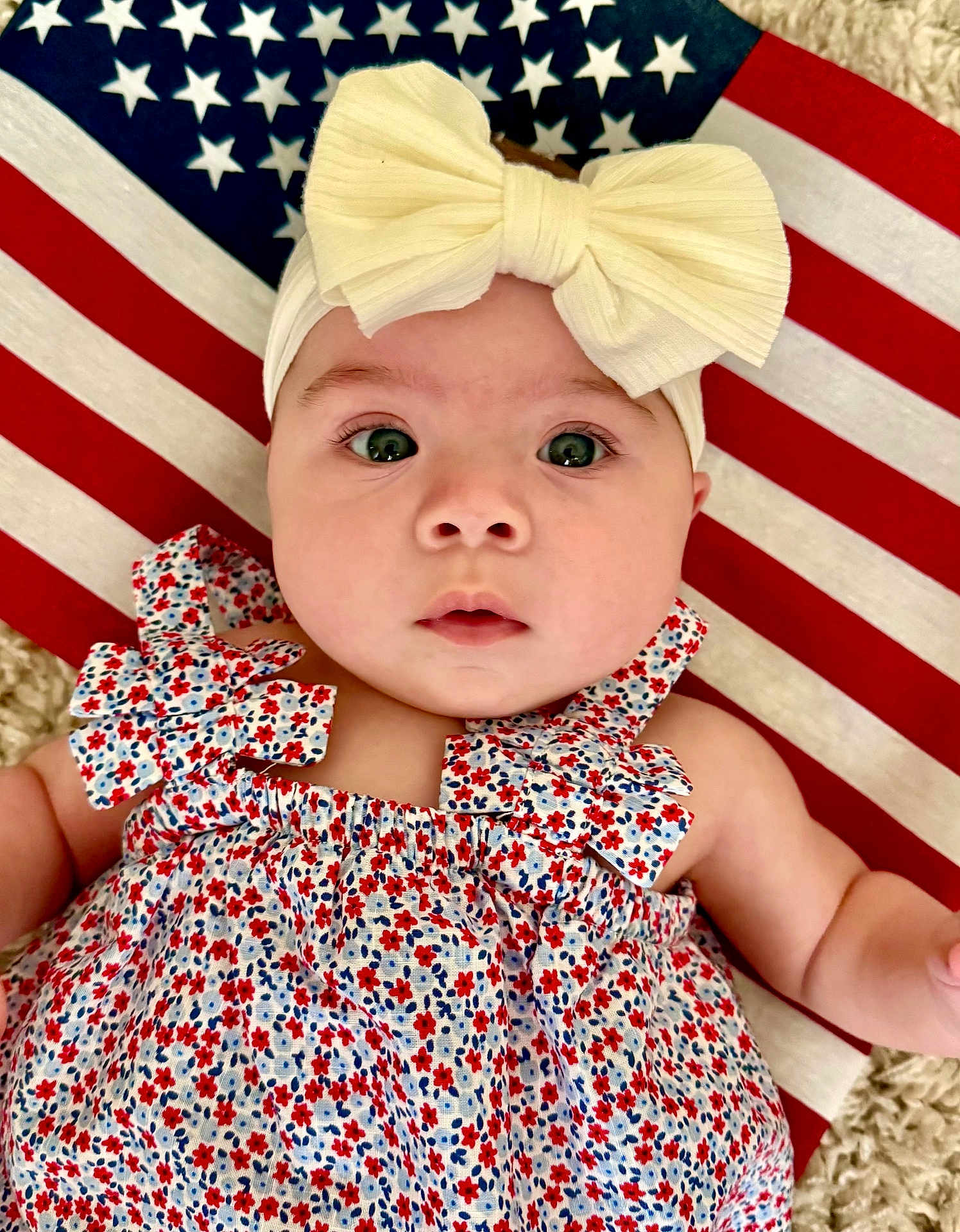 Brenna is registered to the contest to win money with this photo: baby, infant, headband, bow, dress, floral_pattern, american_flag, blanket, face, eyes, skin, arm, cute, child, portrait, laying_down, fabric, pattern, closeup, indoors