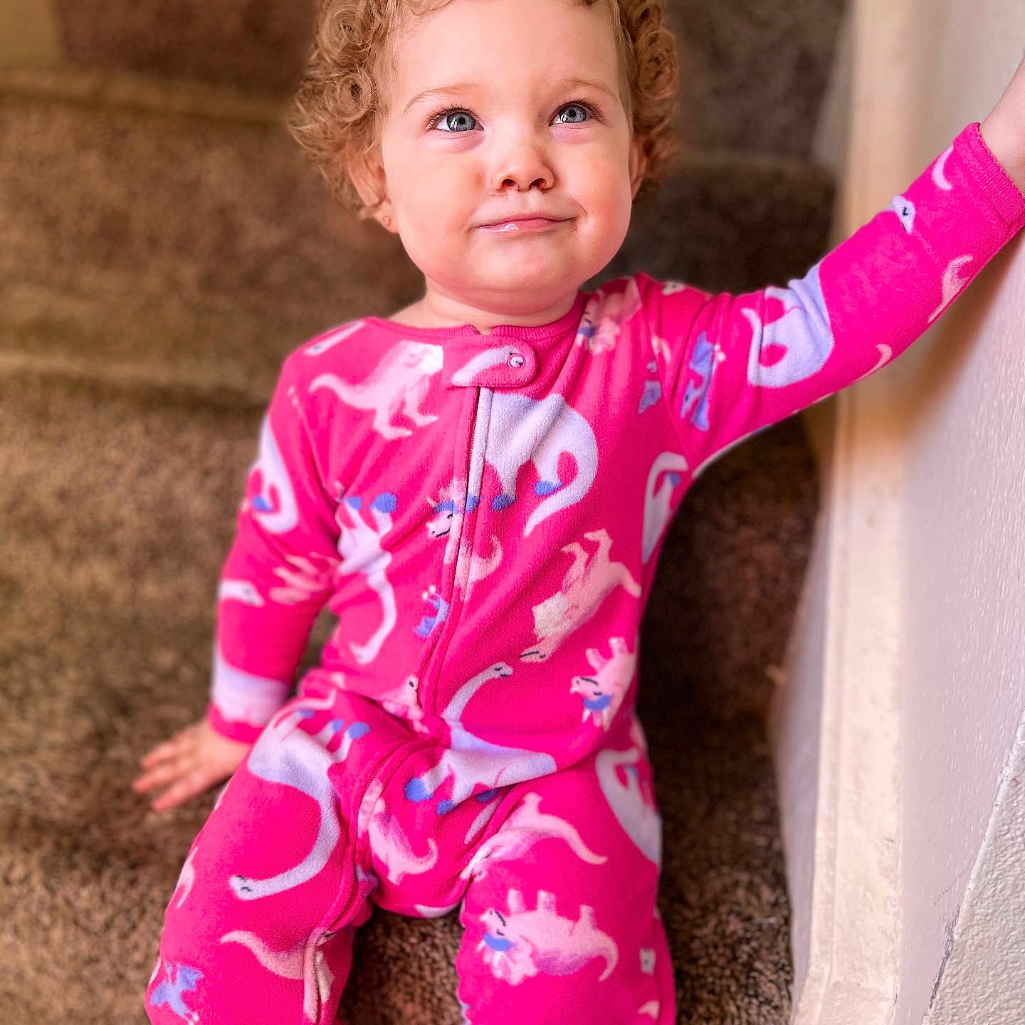 Hollyann joined the competition — help win amazing prizes! blue_eyes, carpeted_stairs, casual_clothing, child, cozy, curly_hair, cute, dinosaur_pattern, hand_on_wall, home, indoor, looking_up, pink_onesie, playful_expression, portrait, seated, smile, stairs, toddler, young_child