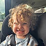 child, toddler, curly_hair, car_seat, seatbelt, smiling, sweater, daisy_pattern, indoors, car_interior, window, sunlight, happy, face, person, cute, portrait, baby, safety, seat