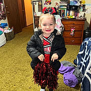 Madison is registered to the contest to win money with this photo: child, girl, cheerleader, pom_poms, black_jacket, white_shoes, carpet, indoor, smile, hair_bow, dresser, books, wall_art, mirror, clothing, toy, room, furniture, happy, person