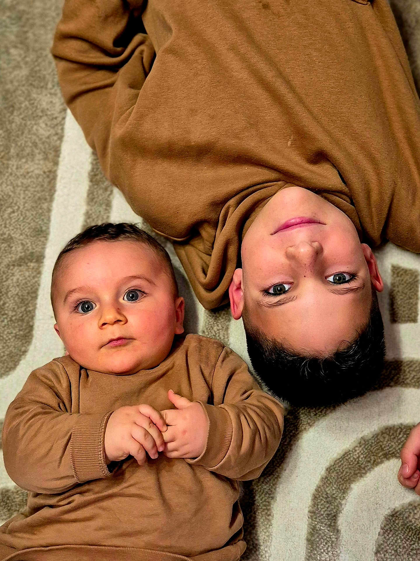 Malone participe au concours pour gagner de l'argent avec cette photo : baby, child, sibling, carpet, brown_clothing, lying_down, indoor, portrait, cute, smile, face, hands, young_child, patterned_carpet, cozy, matching_outfits, curious, eyes, candid, family